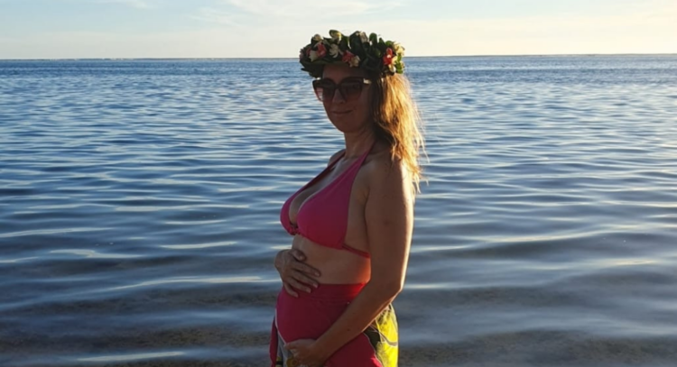 Emeline est en maillot de bain avec une couronne de fleurs sur la tête et caresse son ventre arrondi au bord de la mer.