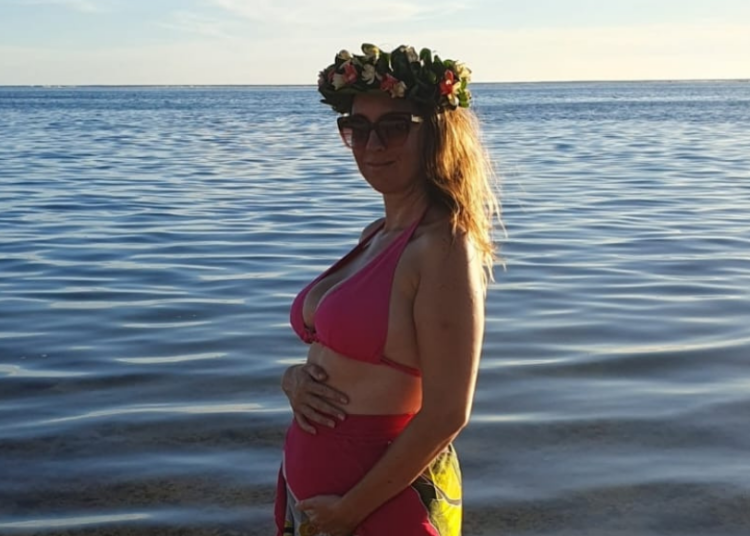 Emeline est en maillot de bain avec une couronne de fleurs sur la tête et caresse son ventre arrondi au bord de la mer.